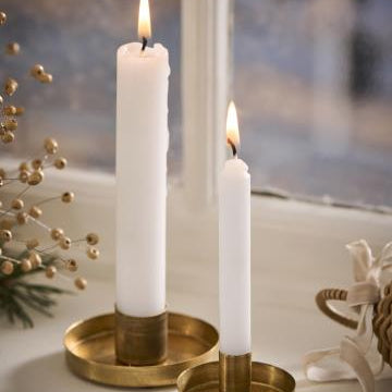Two white candles in brass candle holders on a windowsill with decorative elements.