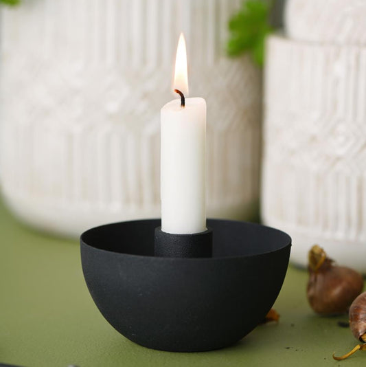 Black candle holder with a lit white candle on a green surface, with plants and decorative items in the background.