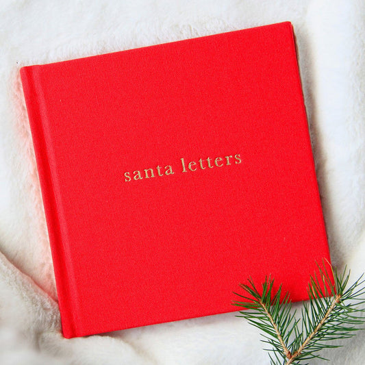 A red keepsake journal with 'Santa Letters' embossed on the cover, placed on a white furry surface with a pine branch to the side.